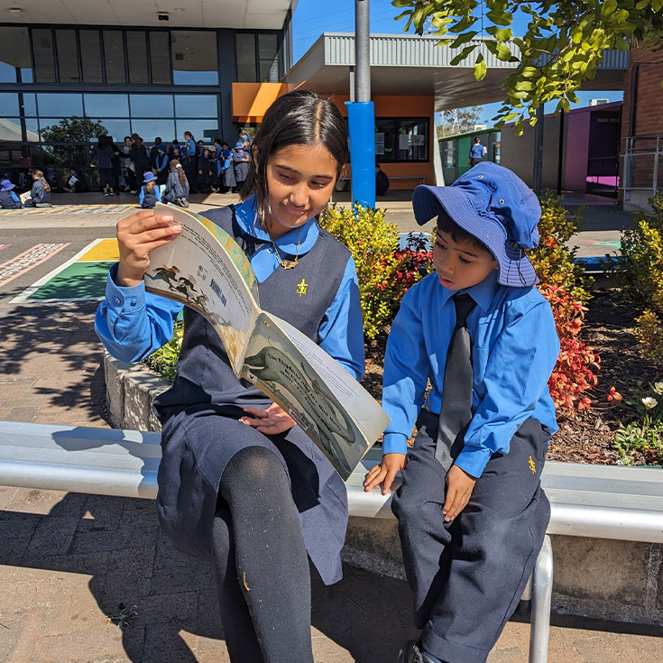 Senior student reading with a kindergarten student in the playground at St Michael's Primary School Blacktown South
